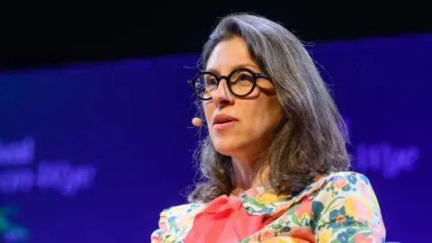Sam Hardwick/Hay Festival A close up image of a woman with dark hair, wearing a multicoloured floral dress and glasses. 