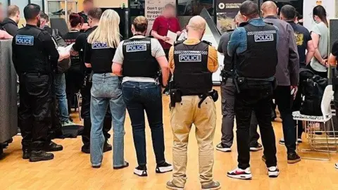 Police officers, some in uniform and some plain-clothed, all wearing stab vests, are briefed in a room with a wooden floor.