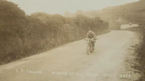 A sepia image from 1907 of the first TT race - a man is on an early motorbike heading from right to left down a road lined with a verge.