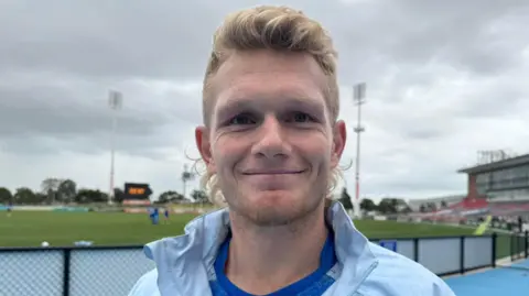 Adam Trealor smiles in a close-up. He has a blond mullet and wears a pale blue sports jacket and royal blue sports shirt. There is a sports field behind him.