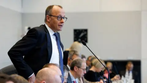 Friedrich Merz stands in front of a microphone wearing a suit and tie to talk to reporters