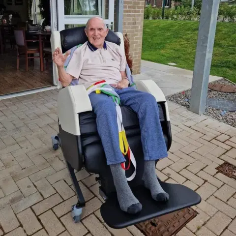 Jack, a smiling man sitting in a reclining wheelchair outside a care home, waving at the camera with the open patio doors and garden visible behind him.