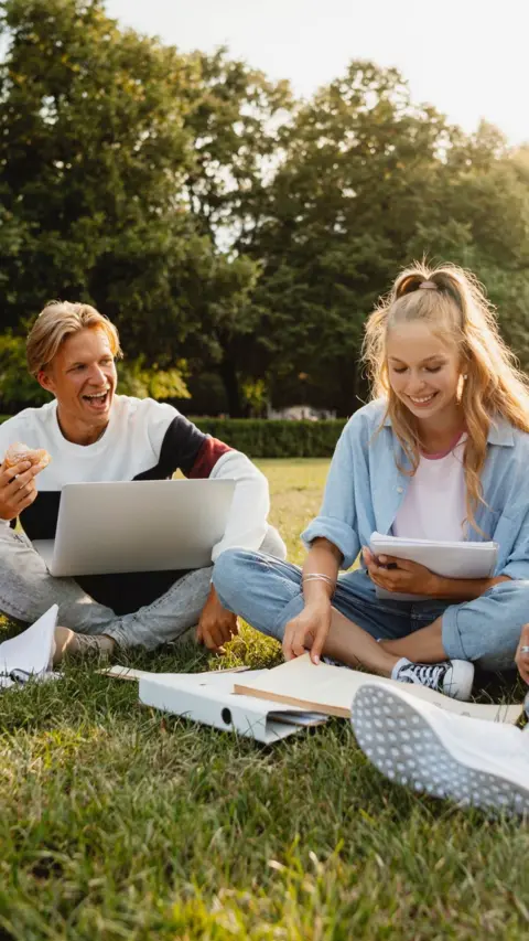 A man with blonde hair in a white jumper and woman with blonde hair in a light blue shirt studying in a park.  