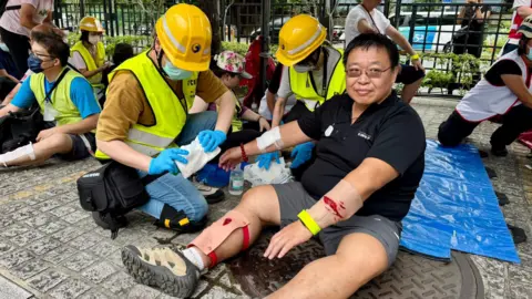 A rescue worker in a yellow helmet and a fluroscent jacket holds a bandage as a volunteer posing as an injured person holds his hand out. The volunteer, dressed in a black t-shirt and grey shorts has fake blood on his right hand and bandages on his left leg and both arms.