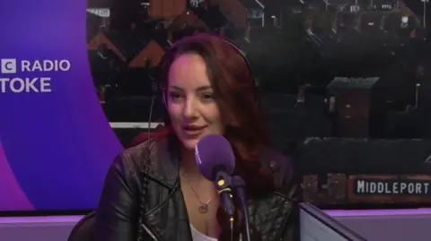 BBC Suzy, a woman sitting in front of a purple microphone, is in a radio studio with a photograph of Middleport Pottery in Stoke-on-Trent behind her. She has dark brown hair and is wearing a black leather jacket.