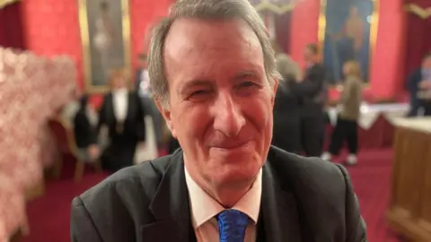 BBC Michael Wills is seen from his shoulders up sitting in a room with a red carpet and red walls. He is wearing a black suit and blue tie and smiling at the camera.