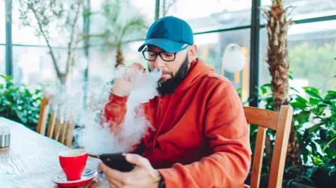 Getty Images Man in red hoody and blue baseball cap blowing a cloud from a vape while looking at his phone with a teacup in front of him