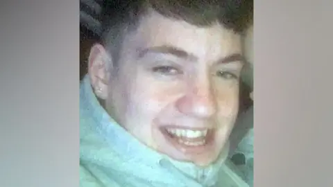 A young man wearing a green jacket and an earring smiles at the camera 