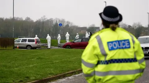 Ant Saddington/BBC Forensics officers dressed in all white with their faces covered standing in the distance on a field. In the foreground, there is a police officer who is looking towards the forensics team. She is wearing a hi-vis yellow police jacket and a black police hat. She is blurred.