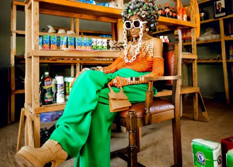 Luxury Media Zambia Margret Chola sits on a wooden chair in what looks like a village shop with produce on the shelves. She wears an afro-shaped hat made of bottle tops, large sunglasses with a white frame and a snow-white chain, green leggings, a sleeveless orange top and matching long gloves, large camel-colored high platforms and holding a small background of the same color