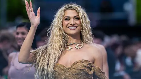 A close-up photo of Antigoni Buxton at the Eurovision opening ceremony in Vienna. She has long, curly blonde hair and is smiling as her right hand is in the air as she waves. She is wearing a gold strapless dress and has gold jewellery around her neck.