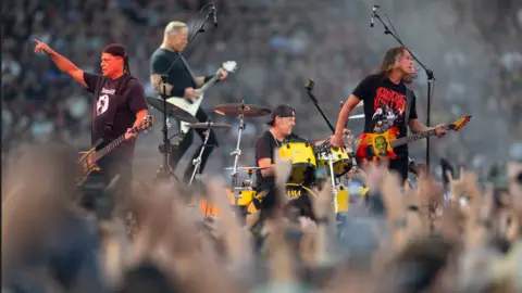 EPA The band play on stage. Raised hands can be seen in the foreground, with seats full of fans seen behind them. A guitarist raises his hand in the air, with the other band members behind him. 