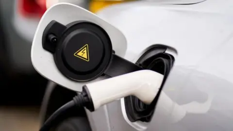 A white car with an electric charging cable plugged into its charging port. The car has its charging port flap open and a yellow electricity warning triangle sticker sits on the inside.