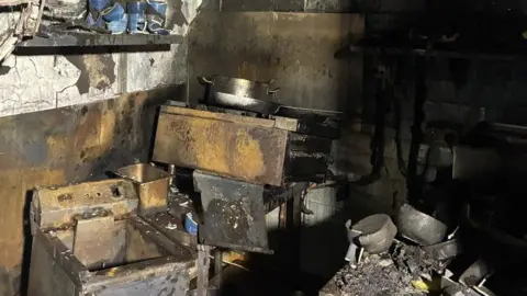 Adam Noble Charred cooking equipment stacked in piles, surrounded by ash.