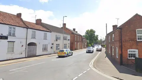 Google A street view of High Street in Gosberton. Red and white period buildings line either side of the main road. A police car is travelling south and a white car is moving in the opposite direction. There are cars parked on the right further down the road.