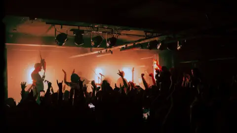 The inside of Independent during a concert. The stage is lit up in orange with multiple band members on stage. A man on the left is holding his guitar above his head. The audience is in shadow but many have their arms in the air.