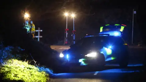 A police car blocks the t-junction at the scene of the crash. It is dark and temporary lights have been set up by emergency workers. The police car has its blue lights flashing. Two people in hi-viz are standing on an embankment looking towards an area of hedge on the corner of the junction.
