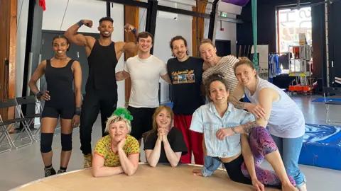 A group of men and women are wearing gym clothes and smiling. They are standing in a studio in front of a circular structure with aerial silks and crash mats in the background. 