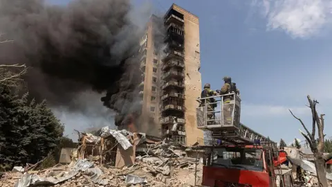 Getty Images A half-ruined building is burning. It is surrounded by brick rubble as black smoke billows out the side of it. A red truck has two firefighters dressed in brown overalls on top of it observing. The sky is clear and blue. A green tree sits to the left of the picture. 