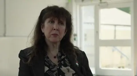 Sharon Coyne is interviewed in a white room. She has long dark hair and is wearing dark clothes.