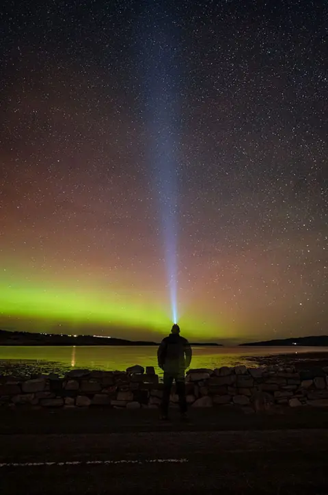 Gary Macleod The Northern Lights in the north coast of Scotland. There are green and red hazes across the sky with Gary Macleod standing on a rocky beach in the foreground. 