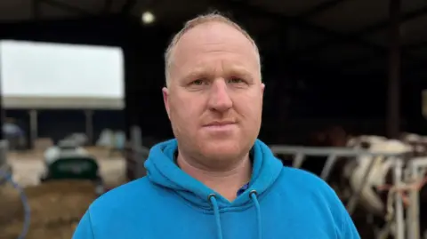 A man, with short light hair, staring at the camera. He's wearing a blue hoodie. 