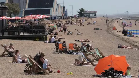 PA Media Dozens of people are seen sat on Weymouth beach, some with parasols and deckchairs, with a beachfront cafe seen in the background