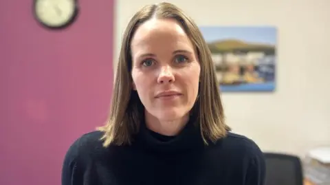 Fiona Hatton has brown shoulder length straight hair and blue eyes. She's wearing a black jumper. She's standing in a room with a purple wall to the left and cream wall with a landscape picture on the wall to the right.