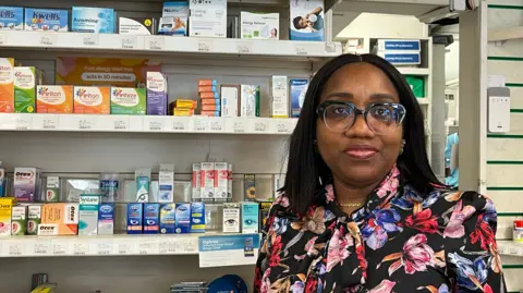 Ifesi Anyamene stands in front of a shelf full of medicines