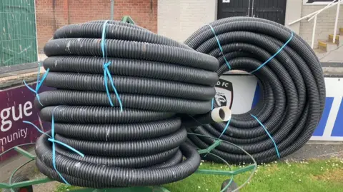 Black drainage tubes are tied up by a football pitch near advertising hoardings. Steps and a wall are in the background.