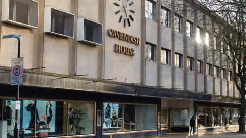 A photo showing the outside of the Cavendish House building in Cheltenham, with a clock and sign and windows below