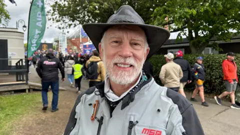 Curt Chapman wears a leather wide-rimmed hat, and a grey bike jacket. He has a white beard. He stands close to the TT grandstand, which is busy with other bike fans walking by.