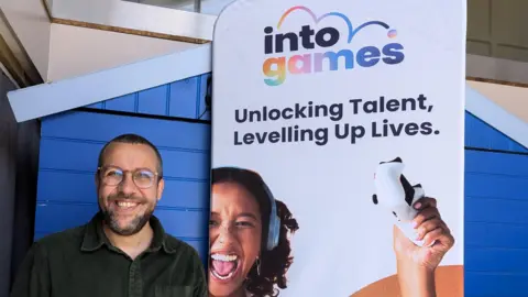 BBC A man smiling and wearing glasses standing next to a banner which reads "Into Games. Unlock Talent, Levelling Up Lives." The woman on the banner is laughing with her mouth open and headphones on, and holding up a controller.