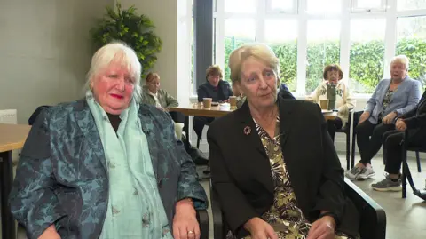 Two sisters sit together in a community hall locaiton. One of them has white hair and is wearing a blue floral suit jacket and light green scarf with bees on it. The other has light brown hair, wearing a black suit jacket and a yellow and green floral dress.