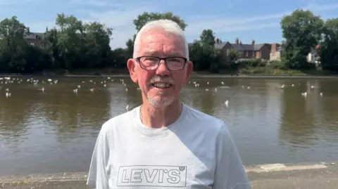 Brian has short grey hair and is wearing a white t-shirt and glasses.