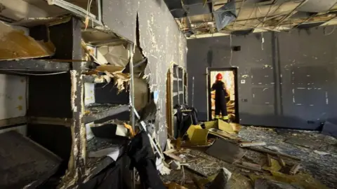 Ministry of Internal Affairs of Ukraine Damaged interior with debris and worker standing in doorway amid wreckage.