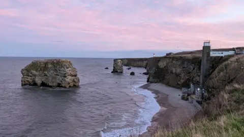 Purple clouds over Marsden Rock, a large rock surrounded by the sea. It is a short way away from a beach, where there is a restaurant at the bottom with a large concrete lift shaft leading to the top