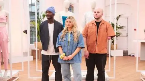 ITV Ryan wearing an orange shirt stood beside two fellow contestants and in front of a collection of white fashion mannequins 