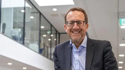 Laurence Frewin photographed at the college. He is seen wearing a grey suit with a light blue shirt. He has stubble and brown swept back hair. He is also wearing glasses and smiling at the camera.