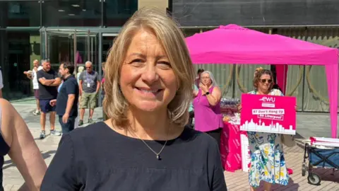 Karen Rose with bobbed blond hair wears a black t-shirt. She stands outside the Plaza building in Liverpool city centre with protesters with a hot pink marquee standing behind her.