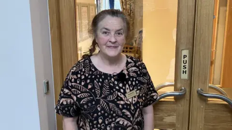 Stephanie Westlake owner of Hummingbird Care Home, stands in front of a pair of double doors with wooden frames, metal handles and clear glass panels. She is wearing a black and white top with patterns made of floral print
