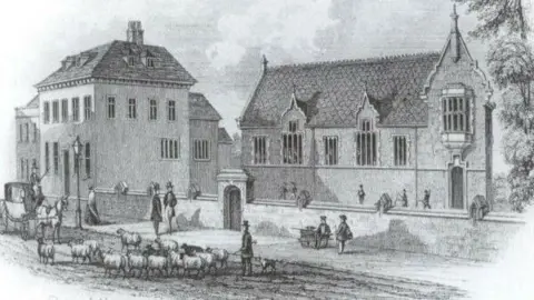 Lichfield District Council A black and white historical image of the building. The date is not known but there are sheep in the road and a horse and carriage, and men are walking on the pavement with top hats and canes with boys from the school nearby.