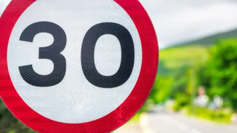 A close up of a 30mph road sign on a sunny day, with trees in soft focus behind it.
