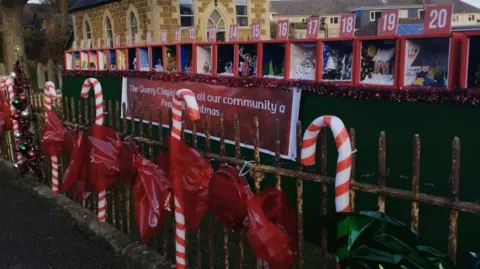 A picture of the advent calendar lined up on a wall. There are red boxes with glass at the front. Inside them are different Christmas themed arts and crafts. On the fence there are candy canes and sweets made from bright red film.