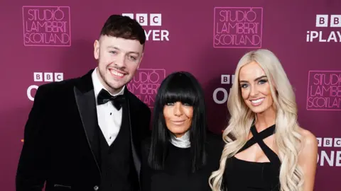 PA Media The Traitors series three winners Jake Brown and Leanne Quigley, pictured with presenter Claudia Winkleman, wearing black tie 