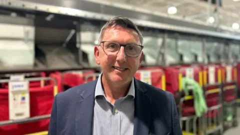 A man with grey hair and black framed glasses, with a navy blazer and striped shirt, smiles at the camera. Behind him is a row of red parcel collection bins under a silver conveyor belt system.