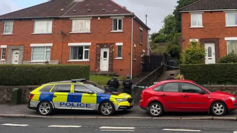 Red brick semi-detached houses. A police cordon is in place at the drive on a house at the right hand side of the picture. In the foreground is a police car and three police officers are standing outside the cordon. There is also a red car in front of the house with the cordon.