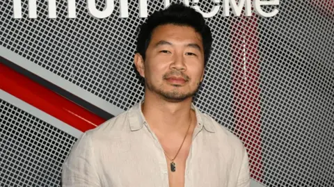 Getty Images Actor Simu Liu smiles at the camera. He is wearing a beige shirt and a gold necklace.