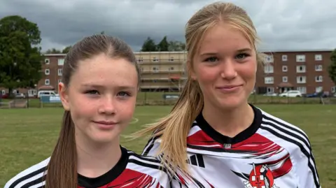 Two girls in Crusaders FC away shirts (white with red and black markings) stand on a football pitch. Low-rise flats and trees are in the background. Rachael has a long brown ponytail and is shorter than Daisy who has a long blonde ponytail.
