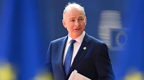 Getty Images Martin wearing a navy suit jacket, blue tie and white shirt. He is looking to the side and speaking. The background is mainly bright blue.